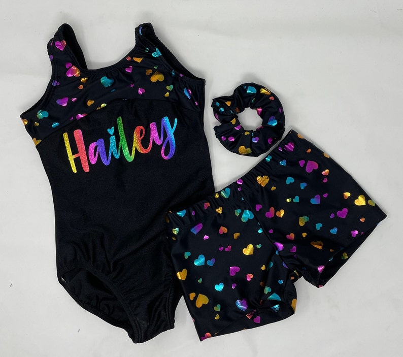 May include: A black leotard, shorts, and scrunchie set with a rainbow heart pattern. The leotard has the name "Hailey" in rainbow lettering. The set is designed for gymnastics or dance.
