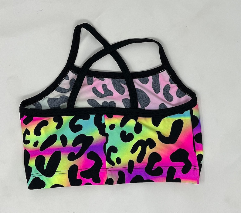 K&ouml;nnte beinhalten: Ein farbenfrohes Crop-Top mit einem lebendigen Leopardenmuster. Das Oberteil hat einen Regenbogen-Farbverlauf aus Neonfarben, darunter Pink, Gelb und Lila, mit schwarzen Leopardenflecken. Die Tr&auml;ger und der Besatz sind schwarz.