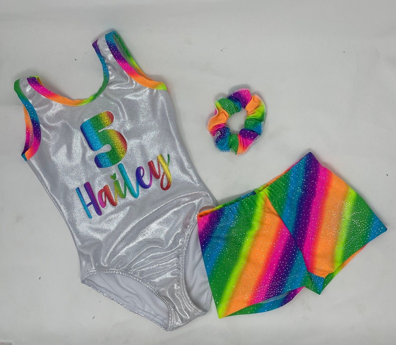 May include: A silver gymnastics leotard, shorts, and scrunchie set. The leotard has a rainbow number 5 and the name "Hailey" in rainbow lettering. The shorts and scrunchie feature a rainbow stripe design.