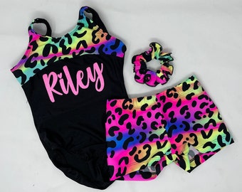 Peut inclure: Ensemble de justaucorps, short et chouchou noirs. Le justaucorps présente un imprimé léopard coloré sur le haut et le nom "Riley" en rose. Le short et le chouchou arborent le même motif léopard. L'ensemble est conçu pour la danse ou la gymnastique.
