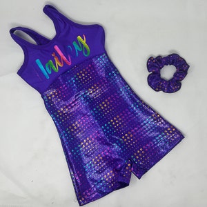 May include: A purple gymnastics leotard with the name "lailey" in rainbow lettering. The shorts portion has a shiny, dotted pattern in various colors. A matching purple scrunchie with gold dots is also included.