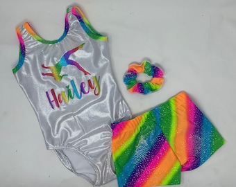 Peut inclure: Ensemble de gymnastique argenté comprenant un justaucorps, un short et un chouchou. Le justaucorps présente un motif de gymnaste arc-en-ciel et le nom "Hailey". Le short et le chouchou présentent un motif à rayures arc-en-ciel. L'ensemble est conçu pour la gymnastique.