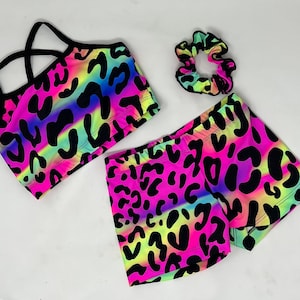 K&ouml;nnte beinhalten: Ein neonfarbenes Sport-Set mit Sport-BH, Shorts und Scrunchie. Das Set hat einen lebendigen Leoparden-Print in Pink, Gelb und Blau mit schwarzen Flecken. Der Sport-BH hat schwarze Tr&auml;ger, und die Shorts sind kurz.
