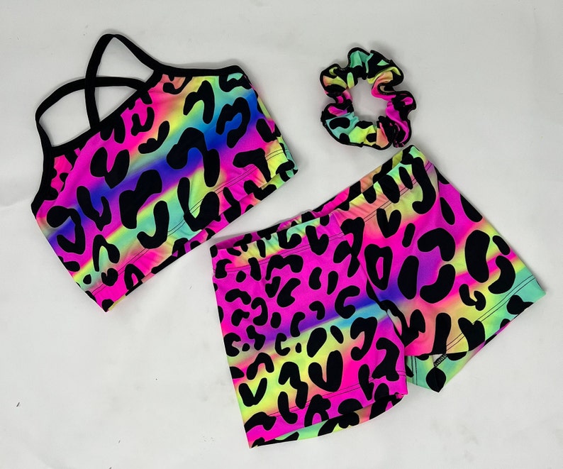 K&ouml;nnte beinhalten: Ein neonfarbenes Sport-Set mit Sport-BH, Shorts und Scrunchie. Das Set hat einen lebendigen Leoparden-Print in Pink, Gelb und Blau mit schwarzen Flecken. Der Sport-BH hat schwarze Tr&auml;ger, und die Shorts sind kurz.
