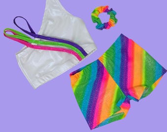 Könnte beinhalten: Ein weißes One-Shoulder-Bustier mit drei diagonalen Trägern in Lila, Grün und Pink, kombiniert mit einer regenbogenfarbenen Shorts mit Glitzereffekt und einem passenden Haargummi. Das Set besteht aus einem glänzenden, dehnbaren Stoff.