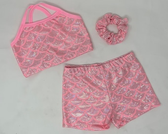 May include: A pink mermaid-scale patterned swimsuit set, including a top with crisscross straps, shorts, and a scrunchie. The set is made of a shiny, iridescent fabric. The swimsuit is designed for children.
