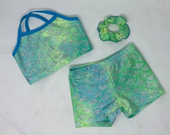 May include: A three-piece set of children's clothing in a mermaid scale pattern. The set includes a crop top with blue straps, matching shorts, and a scrunchie. The fabric has a shimmering effect with shades of blue and green.