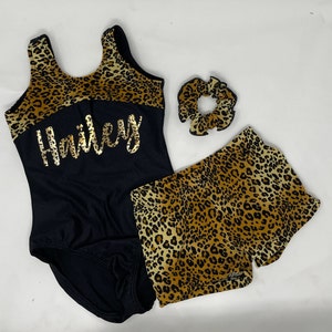Pu&ograve; includere: Un body nero con la parte superiore con stampa leopardata e il nome "Hailey" in lettere dorate. Pantaloncini e scrunchie con stampa leopardata abbinati completano il set. L'outfit &egrave; progettato per la ginnastica o la danza.
