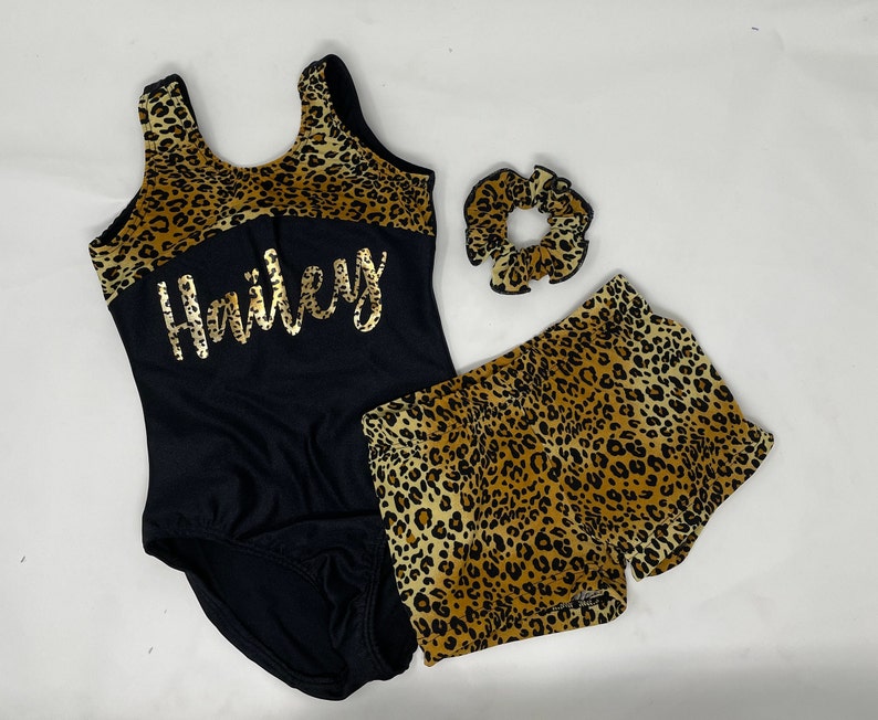 Pu&ograve; includere: Un body nero con la parte superiore con stampa leopardata e il nome "Hailey" in lettere dorate. Pantaloncini e scrunchie con stampa leopardata abbinati completano il set. L'outfit &egrave; progettato per la ginnastica o la danza.