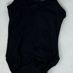 May include: Black sleeveless leotard with a V-neck. The leotard has a scoop back and a fitted shape. The fabric looks like a smooth, stretchy material. The garment is designed for dance or gymnastics.