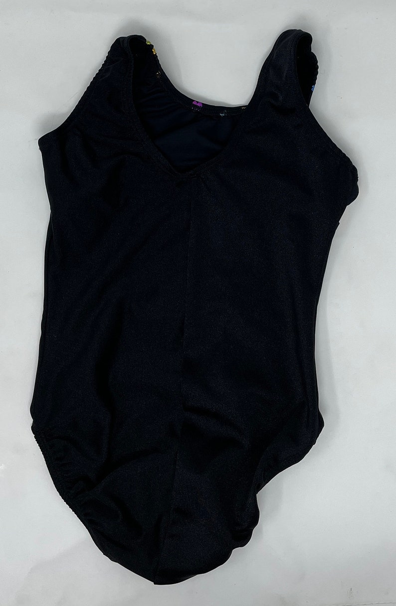 May include: Black sleeveless leotard with a V-neck. The leotard has a scoop back and a fitted shape. The fabric looks like a smooth, stretchy material. The garment is designed for dance or gymnastics.