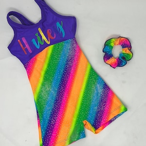 May include: A purple and rainbow-striped leotard with the name "Hiley" in colorful script. The leotard has a scoop neck and a matching rainbow scrunchie. The leotard is covered in small, sparkling embellishments.