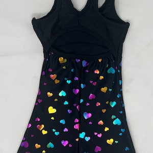 K&ouml;nnte beinhalten: Schwarzer Turnanzug mit offenem R&uuml;cken und Shorts. Die Shorts sind mit einem Muster aus irisierenden Herzen in Gold, Blau, Lila und Rosa bedeckt. Das Oberteil ist einfarbig schwarz mit &auml;rmellosem Design.