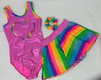 May include: A pink leotard with a rainbow heart design and the name "Haley" in script. A matching rainbow skirt and scrunchie complete the set. The outfit is designed for gymnastics or dance.