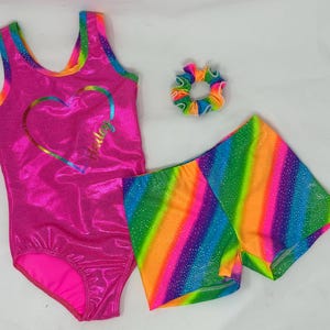 May include: A hot pink leotard with a rainbow heart design and matching rainbow shorts. A rainbow scrunchie is also included. The leotard and shorts have a shiny, glittery finish. The outfit is designed for gymnastics or dance.