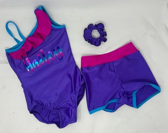 Puede incluir: Un conjunto morado de traje de baño, shorts y coletero. El traje de baño tiene un diseño de un solo hombro con volantes rosas y el nombre "Hailey" en letras iridiscentes. Los shorts y el coletero son morados con detalles rosas y turquesas.