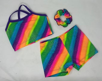 Puede incluir: Un vibrante conjunto de baile a rayas arcoíris, que incluye un top corto, pantalones cortos y una scrunchie. El conjunto presenta un espectro de colores, desde naranja neón hasta morado, con acentos brillantes. El top corto tiene un ribete y correas moradas.
