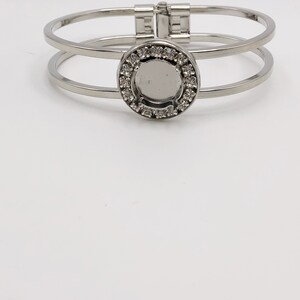 May include: A silver-toned bracelet with a double-band design. The bracelet features a circular centerpiece adorned with small, clear crystals. The clasp is a simple, secure design. The jewelry is set against a white background.