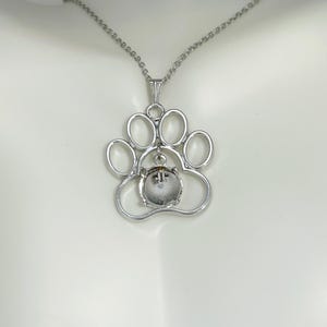May include: A silver-colored pendant necklace featuring a paw print design. The paw print has five open circles and a small, round, clear stone in the center. The necklace chain is also silver and hangs on a white surface.