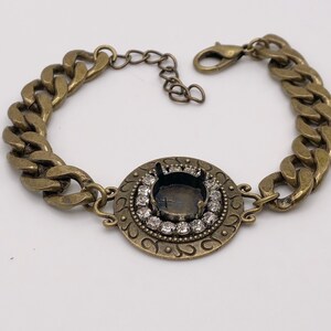 May include: Antique gold-tone bracelet featuring a large, round centerpiece with a setting for a stone, surrounded by small clear crystals. The bracelet has a chunky chain design and a lobster clasp closure.