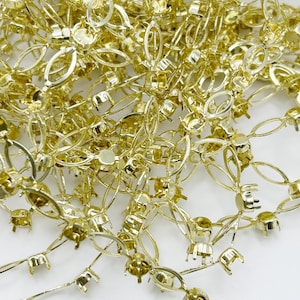 May include: Close-up of a pile of gold-colored metal jewelry findings. The findings have a floral design with oval and circular shapes, and small cup-like settings. The pieces are scattered on a white surface.