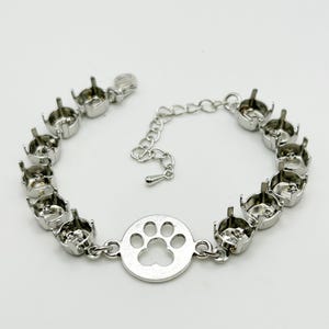 May include: A silver-toned bracelet featuring a paw print charm. The bracelet has multiple settings for stones or gems, and a chain with an adjustable length. The paw print charm is in the center of the bracelet.