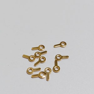May include: A collection of small, gold-colored metal findings, each with a circular loop and a rectangular extension. These findings are scattered on a light gray surface, suggesting their use in jewelry making or crafts.