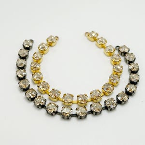 May include: Three delicate necklaces with clear, round gemstones. One necklace has a gold-colored setting, while the other two have a dark gray setting. The necklaces are arranged in a curved shape against a white background.