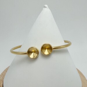 May include: A gold-colored open cuff bracelet. The bracelet features two circular settings on either end, designed to hold a gemstone or other embellishment. The bracelet is displayed on a white, cone-shaped form.