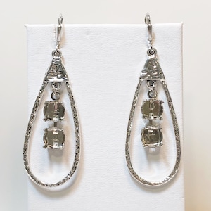 May include: Silver-toned drop earrings with a textured teardrop design. Each earring features three square, light-colored stones. The earrings are displayed on a white surface, showcasing their intricate details and elegant style.