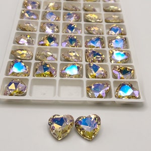 May include: A tray of iridescent crystal rhinestones in various shapes, including teardrops and hearts. The crystals have a shimmering, multi-colored effect, reflecting light with hues of yellow, blue, and purple. Two heart-shaped crystals are placed at the bottom.