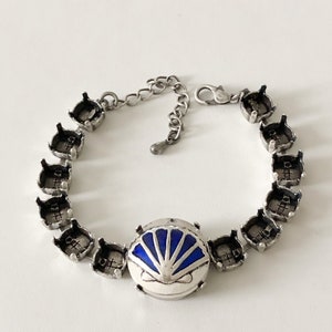 May include: Silver-toned bracelet featuring a central round charm with a blue and white fan design. The bracelet has a series of empty settings around the wrist, connected by a chain with an adjustable clasp. The bracelet is on a white background.