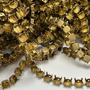 May include: Close-up of a pile of gold-colored metal jewelry findings. The findings are square-shaped with small prongs, designed to hold gemstones. The findings are arranged in a pile and in a line, showing their design and texture.