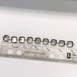 May include: A close-up of eight silver-toned metal settings for rhinestones or other embellishments. The settings are arranged in a row on a ruler with inch and centimeter markings. The ruler shows measurements up to 6 inches or 15 centimeters.