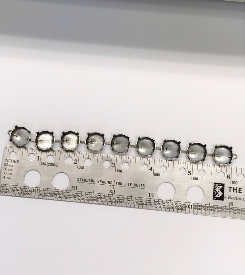 May include: A close-up of eight silver-toned metal settings for rhinestones or other embellishments. The settings are arranged in a row on a ruler with inch and centimeter markings. The ruler shows measurements up to 6 inches or 15 centimeters.