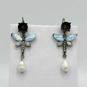 May include: A pair of dragonfly earrings. Each earring features a light blue iridescent wing design, a small cluster of clear crystals, and a dangling white pearl. The earrings have a bronze-colored metal finish and a lever-back closure.
