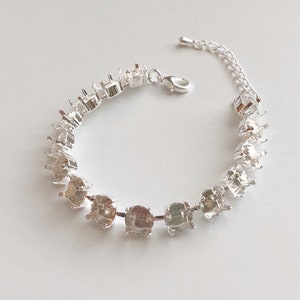 May include: A silver-toned bracelet with multiple square settings for gemstones. The bracelet has a delicate chain and a clasp closure. The settings are empty, ready for customization. The bracelet is displayed on a white surface.