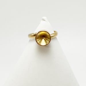 May include: A gold-colored ring with a circular setting. The ring is displayed on a white surface. The setting is a shallow, concave circle, designed to hold a gemstone or other decorative element. The band is smooth and simple.