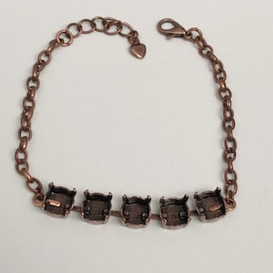 May include: A copper-colored bracelet with a chain design. The bracelet features five square settings for stones or embellishments. The chain includes a heart-shaped charm and a lobster clasp for closure.