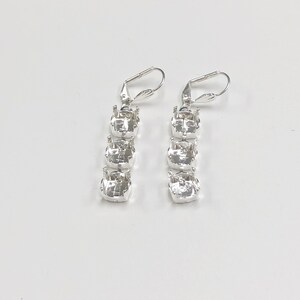 May include: A pair of silver-toned drop earrings. Each earring features three clear, square-cut crystals set in a vertical line. The earrings have lever-back closures and are displayed against a white background.