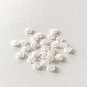 May include: A collection of small, white, round embellishments with a textured, crystal-like appearance. Each piece has iridescent accents, creating a subtle shimmer. These decorative elements are scattered on a white surface.