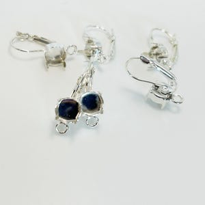 May include: Several silver-toned leverback earring findings with square settings. Two earrings have dark blue, square-shaped stones. The findings are on a white surface.