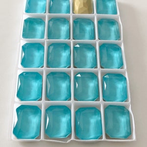 May include: A white plastic tray with 19 compartments, each containing a turquoise blue glass gem. One compartment contains a gold gem.