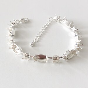 May include: A silver-toned bracelet with multiple settings for gemstones. The bracelet features a series of oval and rectangular settings connected by small links and an adjustable chain. The clasp is a lobster claw style.