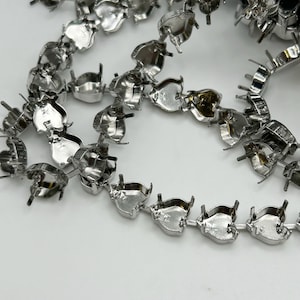 May include: Close-up of multiple strands of silver-colored metal jewelry findings. Each finding is a small, open-backed setting, connected in a chain-like pattern. The findings are arranged on a white surface.