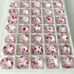 May include: A tray of round, white glass beads with a speckled red pattern. Each bead is nestled in its own square compartment within the white plastic tray. The beads have a smooth, glossy finish and are evenly spaced.