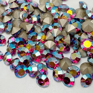 May include: A collection of round, faceted crystals featuring an iridescent finish with vibrant pink, blue, and yellow hues. Each crystal has a pointed, metallic-toned back and a reflective, multi-colored top surface, scattered across a plain surface.