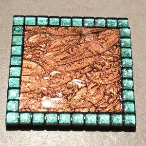 Könnte beinhalten: Quadratische Mosaikfliese mit kupferfarbenem Zentrum und einem Rand aus kleinen, quadratischen, türkis farbenen Fliesen. Das kupferfarbene Zentrum hat ein strukturiertes, organisches Aussehen. Der türkisfarbene Rand fügt eine Kontrastfarbe und ein geometrisches Element hinzu.