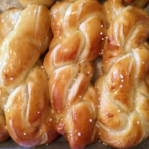 May include: Close-up of several golden-brown, braided bread loaves. The bread has a glossy finish and is sprinkled with coarse salt crystals. The baked goods are arranged in a baking pan, suggesting a fresh-baked theme.