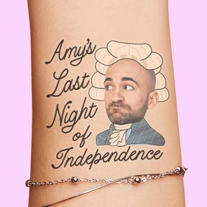 May include: A humorous tattoo on an arm features the text "Amy's Last Night of Independence" in elegant script. The design includes a cartoon illustration of a man in a powdered wig and colonial attire, set against a pink background.